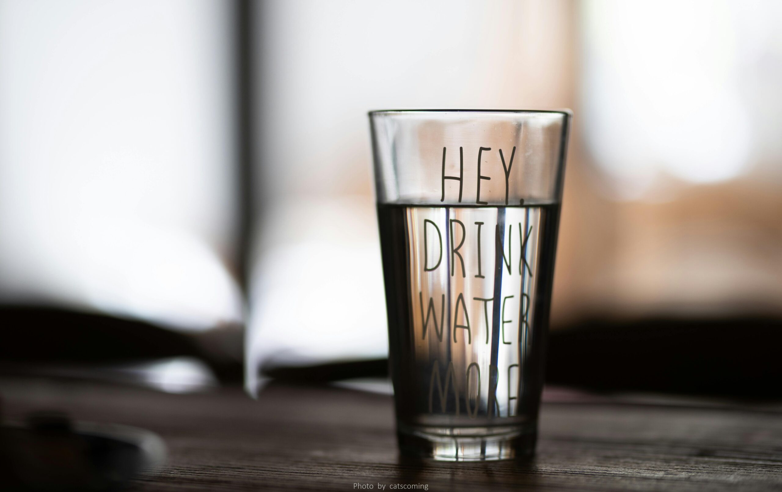 Close-up of a water glass with motivational text 'Hey Drink Water More' in a blurred indoor setting.