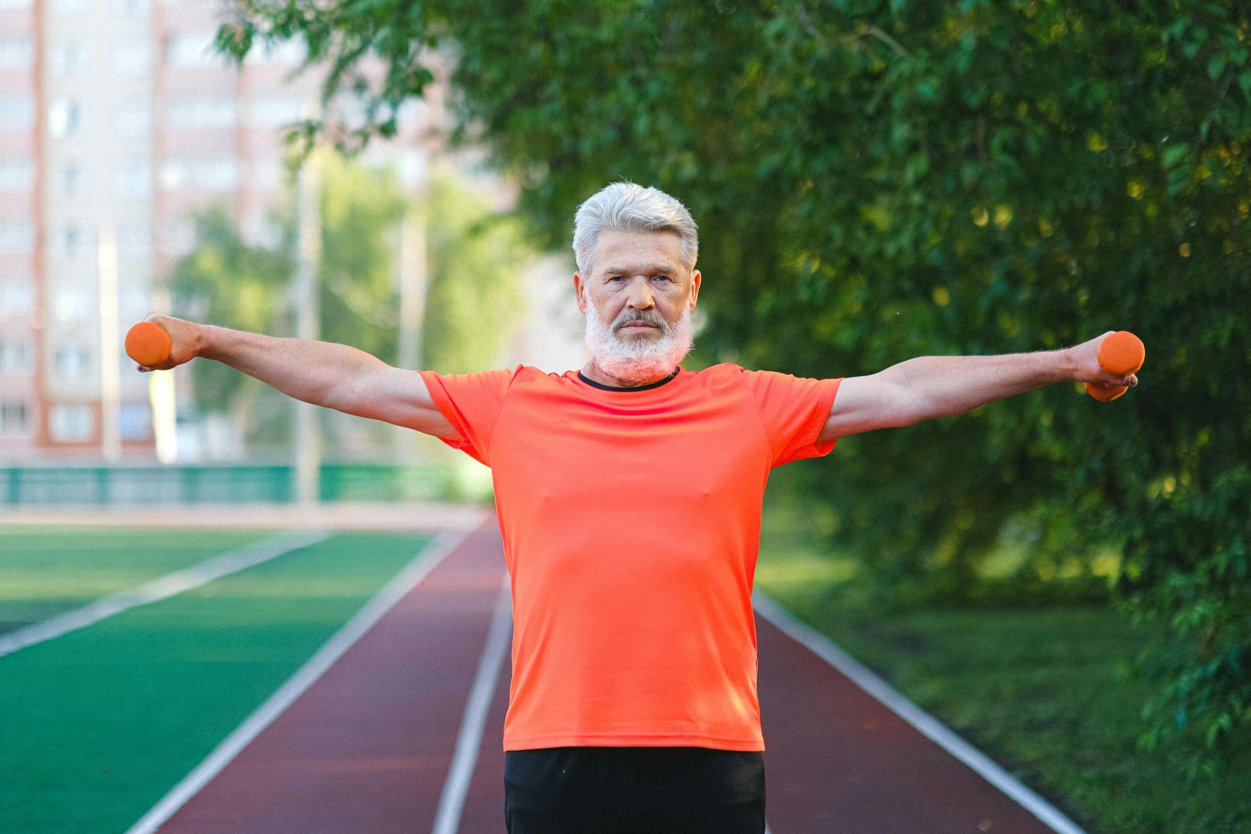 Elderly man in orange shirt exercising with dumbbells on a running track outdoors.