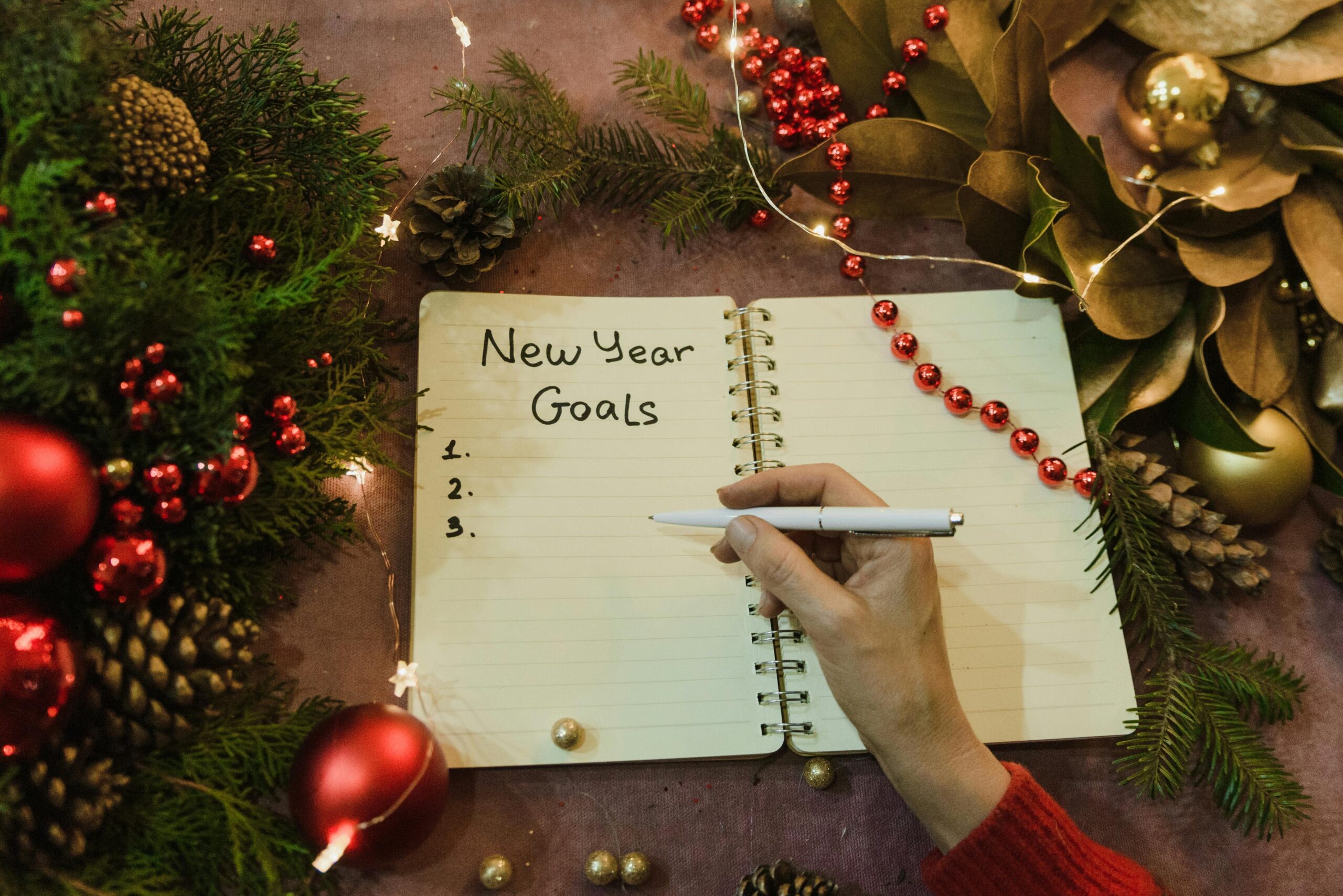 Hand writing New Year goals in a spiral notebook surrounded by festive Christmas decorations.