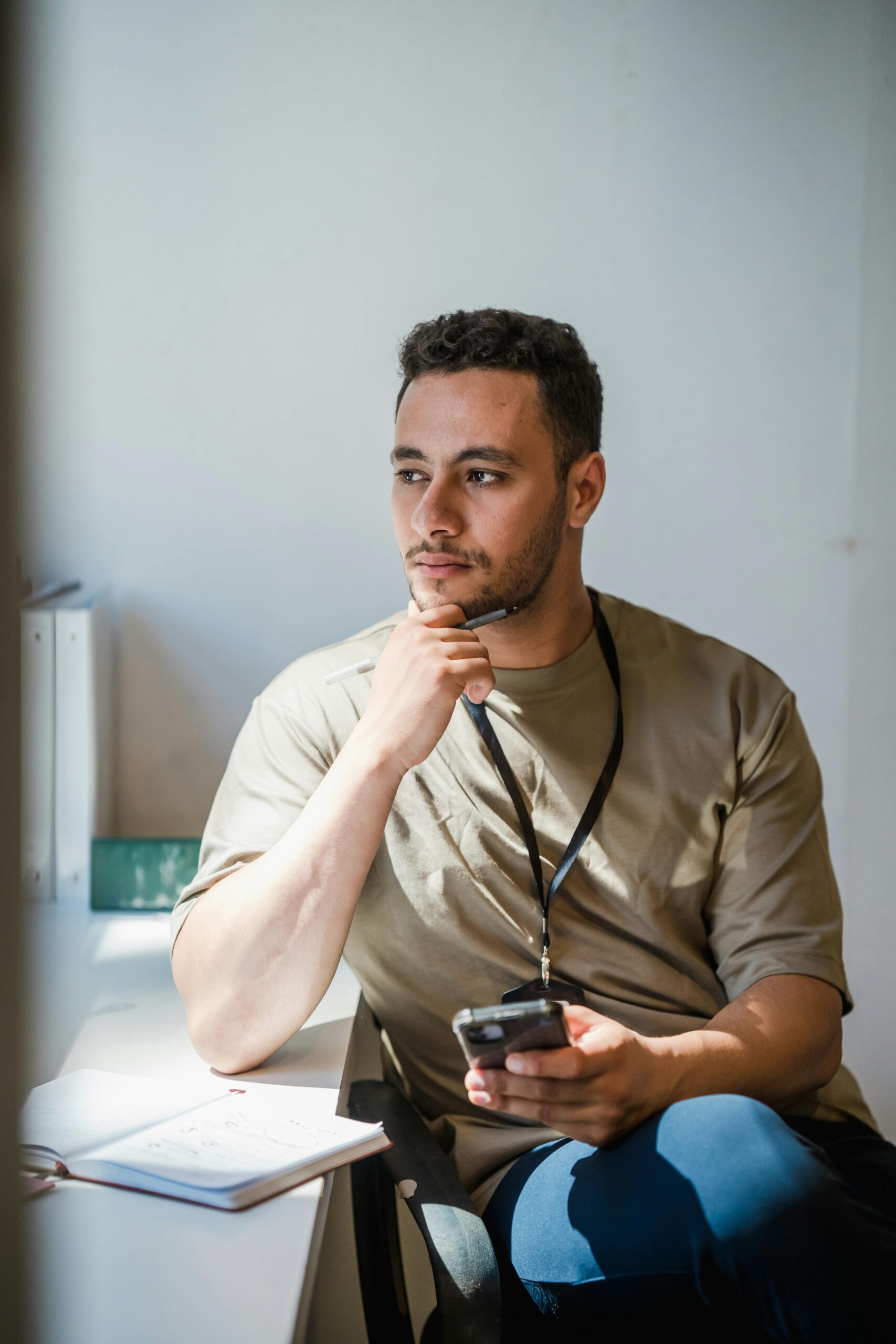 Portrait of a contemplative man sitting by the window in an office, holding a smartphone and thinking.