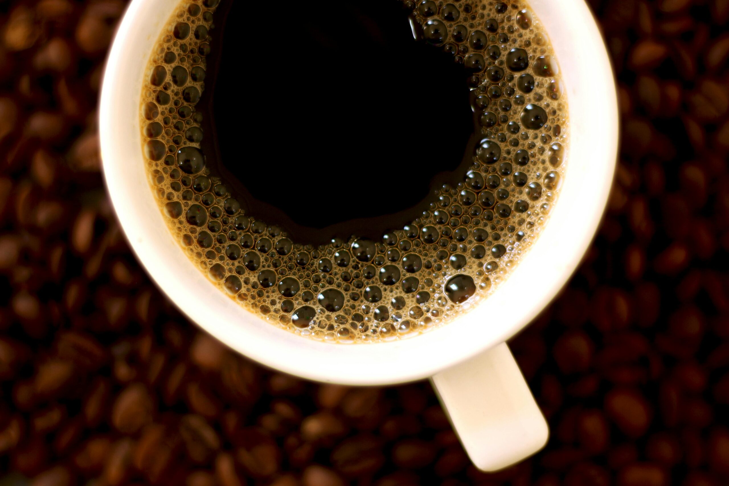 High-angle view of hot black coffee with bubbles in a white porcelain cup over coffee beans.